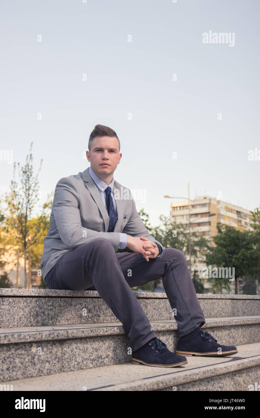 Un giovane uomo caucasico, vuota espressione, Business Suit, formale, usura ordinaria persona comune ritratto, all'aperto seduti sulle scale passi Foto Stock