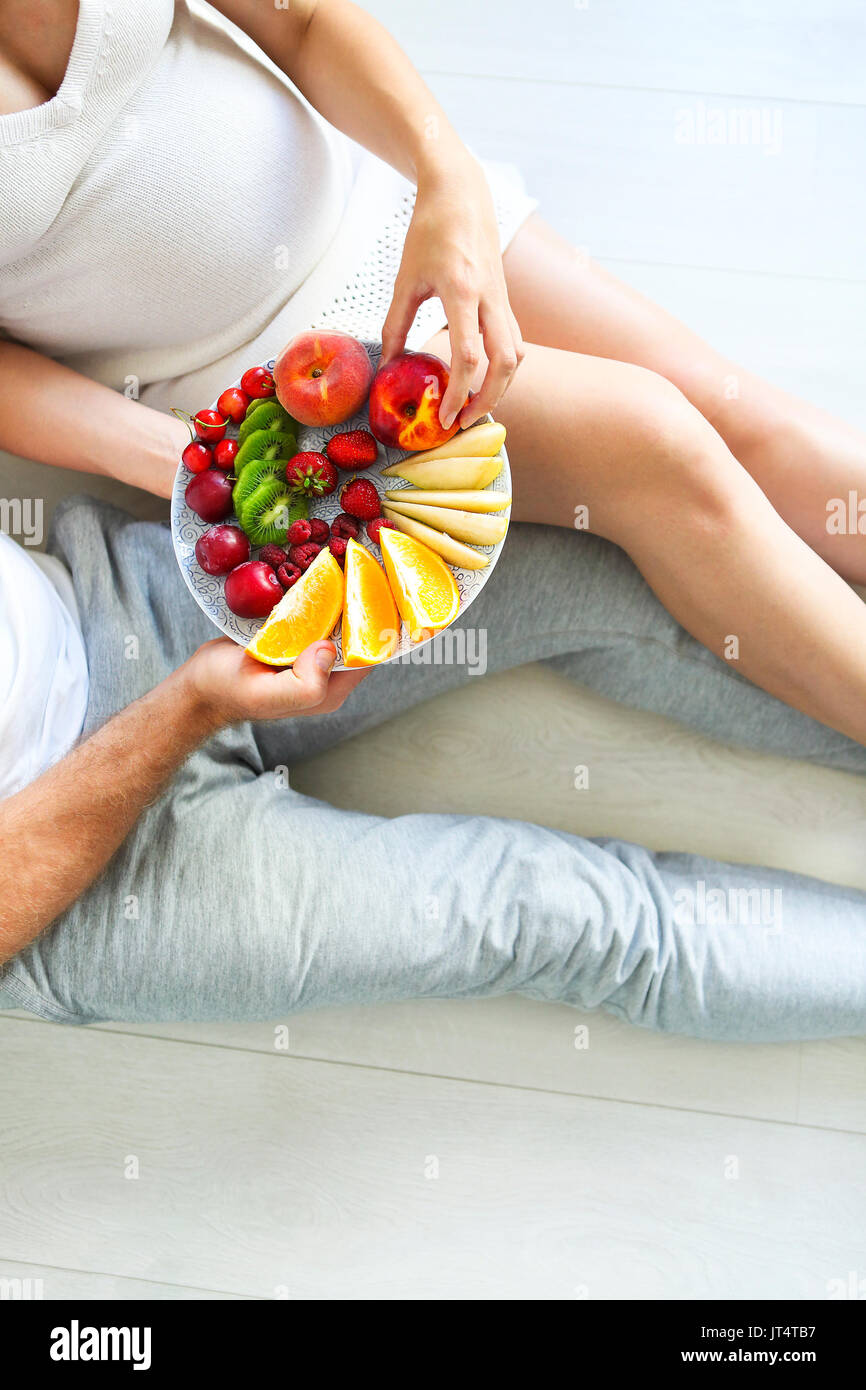 Giovane uomo e donna incinta seduta con un piatto di frutta. La gravidanza e il concetto di nutrizione Foto Stock