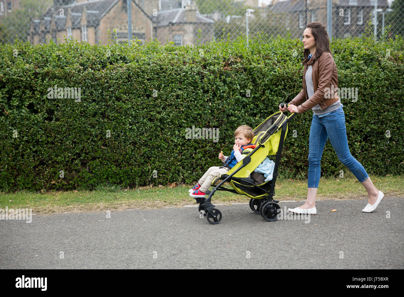 Vista laterale di un Caucasian mamma a camminare su una strada di città mentre si spinge il suo bambino seduto in una carrozzina. Foto Stock