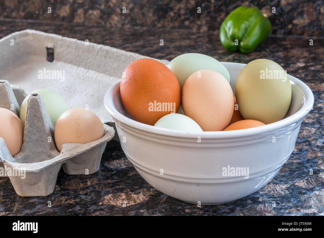 Foto orizzontale multicolore di uova in una ciotola bianco e alcuni ancora nella scatola di cartone grigio a lato su un banco di cucina con un peperone verde Foto Stock