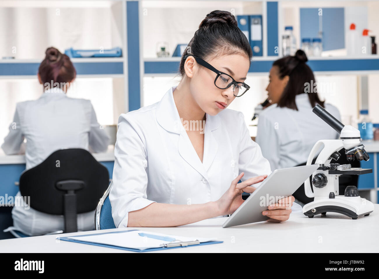 Scienziato asiatico in camice da laboratorio con microscopio e tavoletta digitale lavorando nel laboratorio chimico, scienziati gruppo dietro Foto Stock