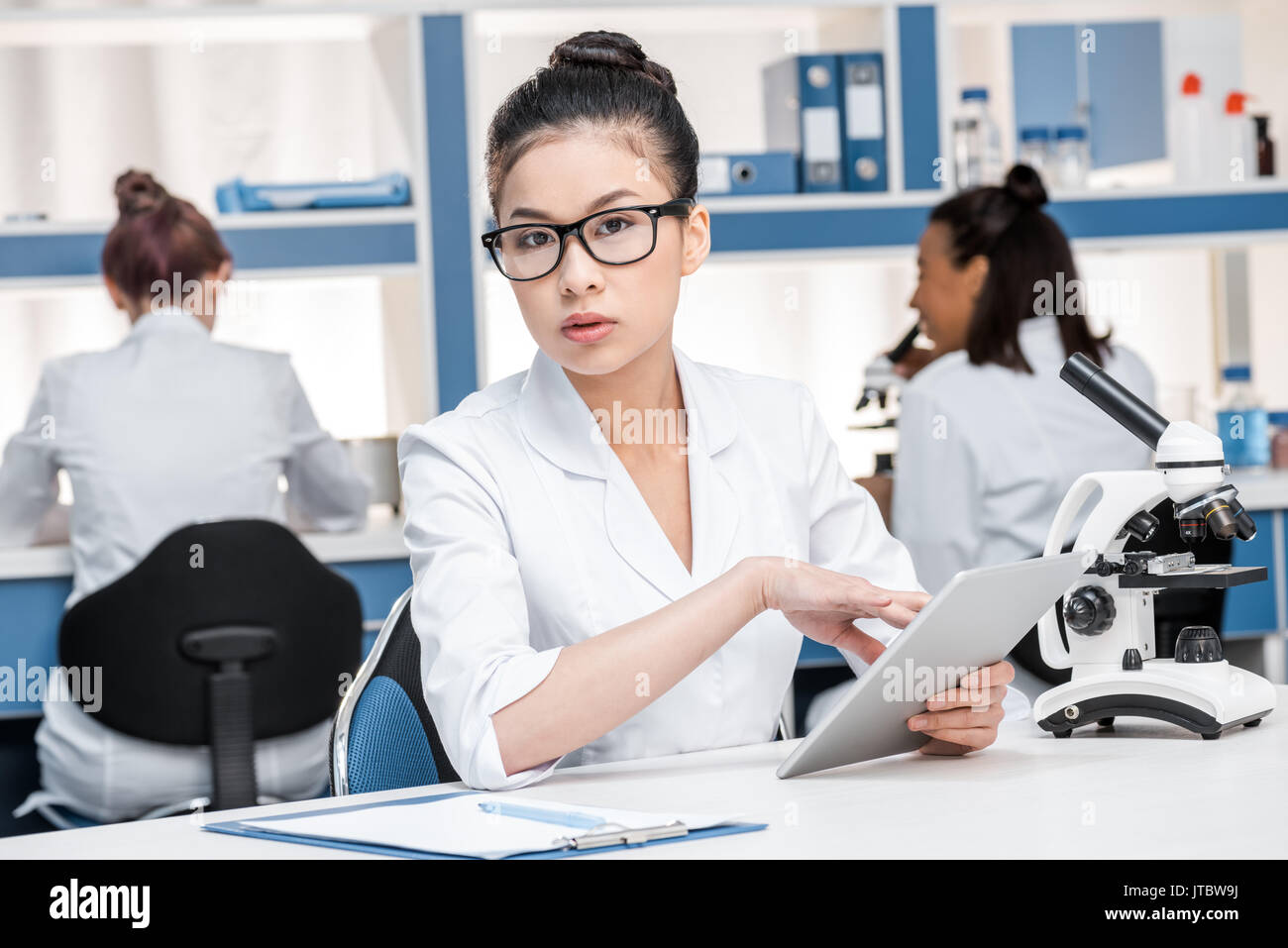 Scienziato asiatico in camice da laboratorio con microscopio e tavoletta digitale lavorando nel laboratorio chimico, scienziati gruppo dietro Foto Stock