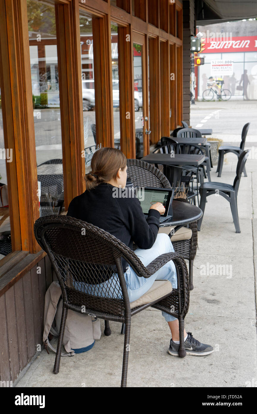 Giovane donna seduta al tavolo al di fuori di un cafe utilizzando una compressa, Main Street, Vancouver, BC, Canada Foto Stock