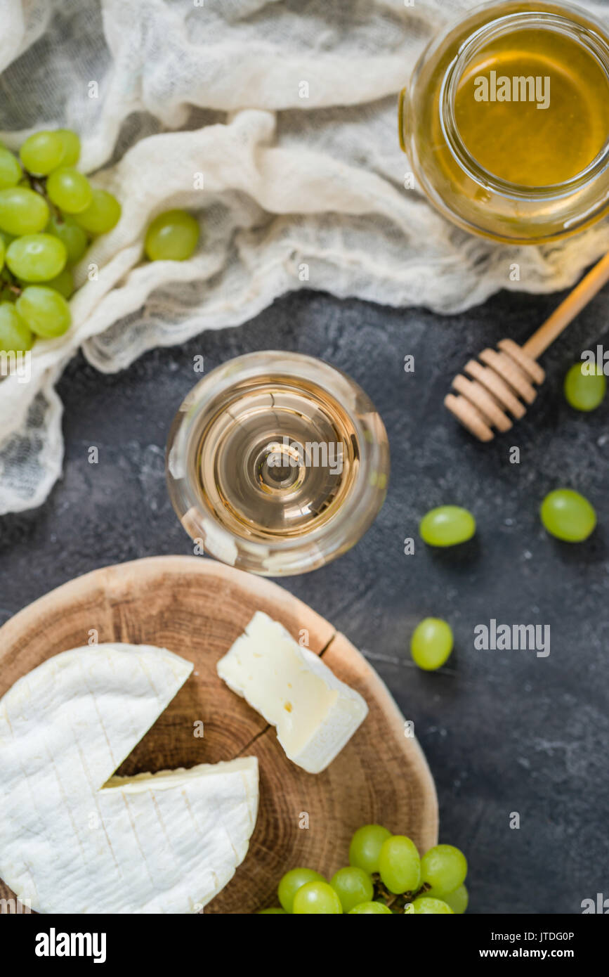 Francese di formaggio morbido dalla regione della Normandia affettato su un taglio di legno verde con uva, miele, cucchiaio e bicchiere di vino bianco sul buio tavolo rustico, offuscata b Foto Stock