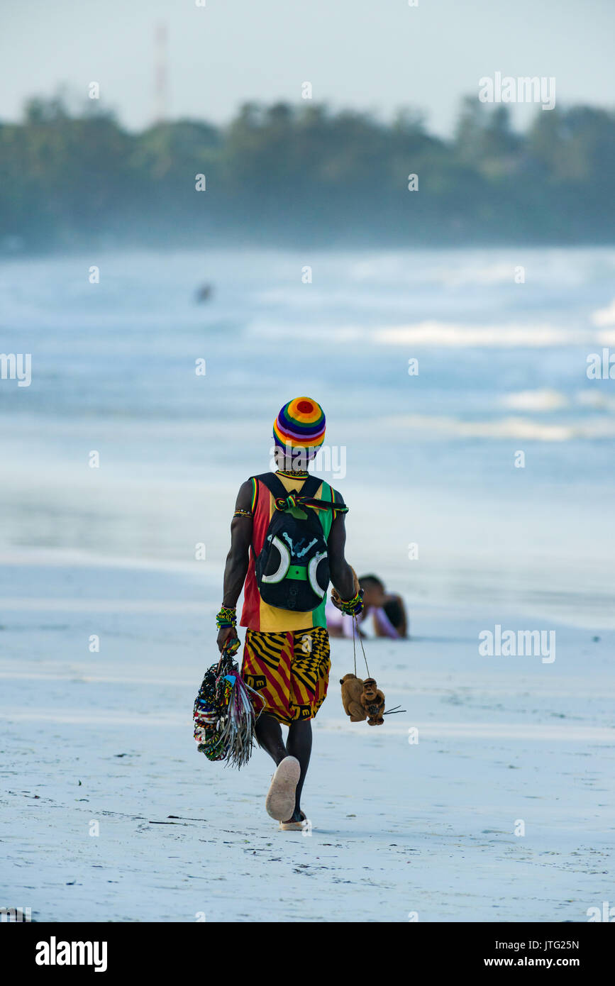 Uomo locale passeggiate lungo la spiaggia con prodotti per la vendita in tarda serata luce, Diani, Kenya Foto Stock