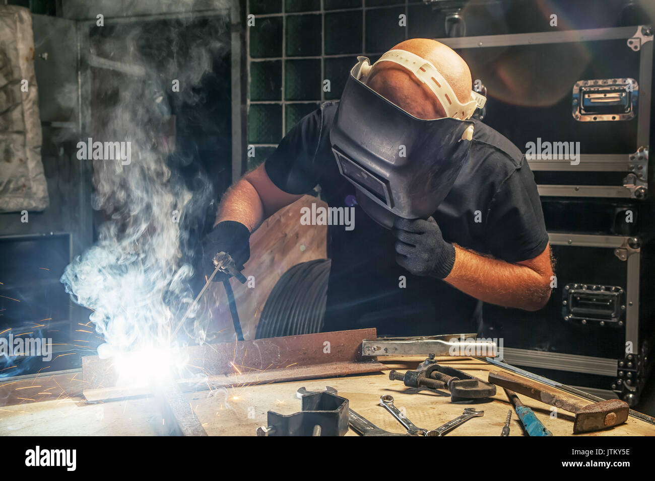 Un giovane uomo calvo in una saldatura maschera nera e black t-shirt infusi un arco di metallo di saldatura la costruzione della macchina in un workshop scuro con una verde parete in vetro Foto Stock