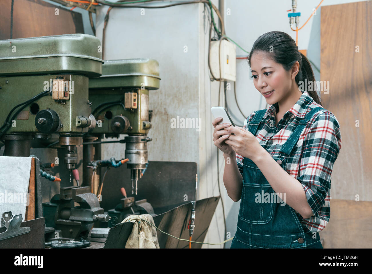 Sorridenti milling machine company personale femminile lavora nel reparto di foratura e utilizzando mobile smart phone per rilassarsi durante il resto del tempo. Foto Stock