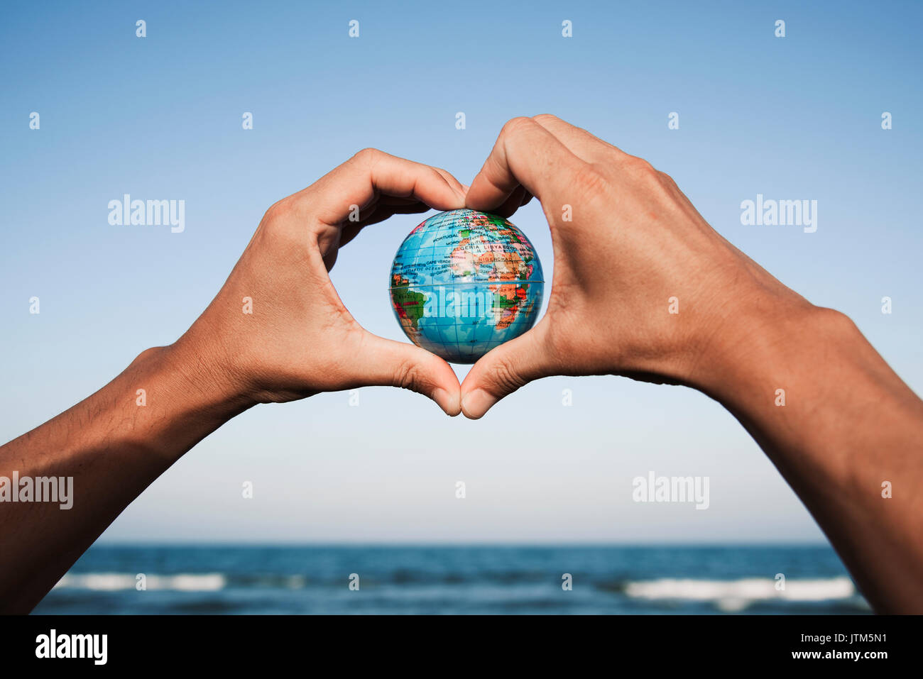 Primo piano di un giovane uomo caucasico con un globo mondo nelle sue mani formando un cuore contro il cielo blu, di fronte al mare Foto Stock