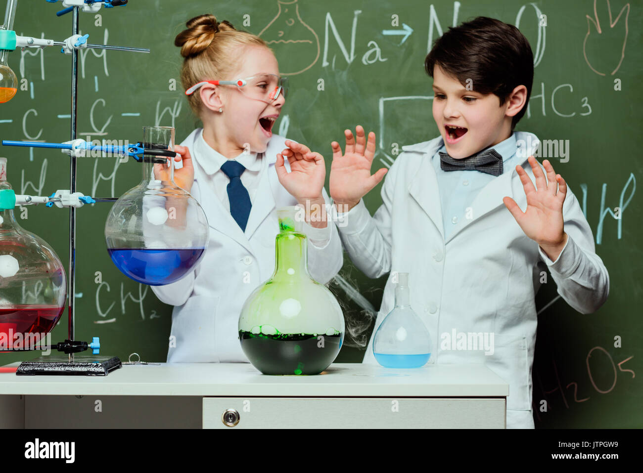 Bambini piccoli in camici con lavagna dietro in laboratorio, scienziati kids concetto del team Foto Stock