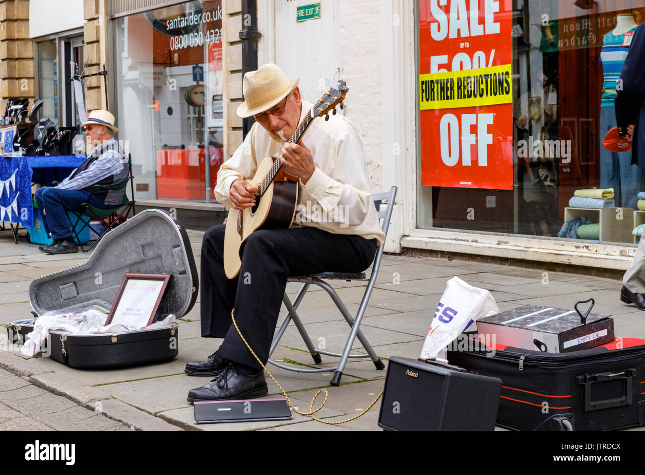 Melton Mowbray folk festival e la fiera artigianale, senior musicista di strada seduto a suonare la chitarra al di fuori della banca di Santander a fianco di un commerciante di mercato Foto Stock