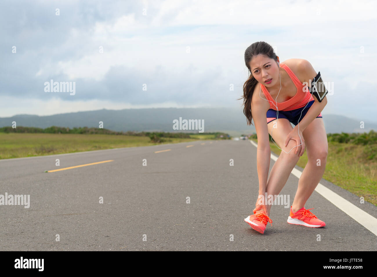 Giovane bella jogging in esecuzione su strada troppo stanco portando la caviglia e il ginocchio dolore scomodo, mostrando l'espressione dolorosa con paesaggio di montagna. Foto Stock