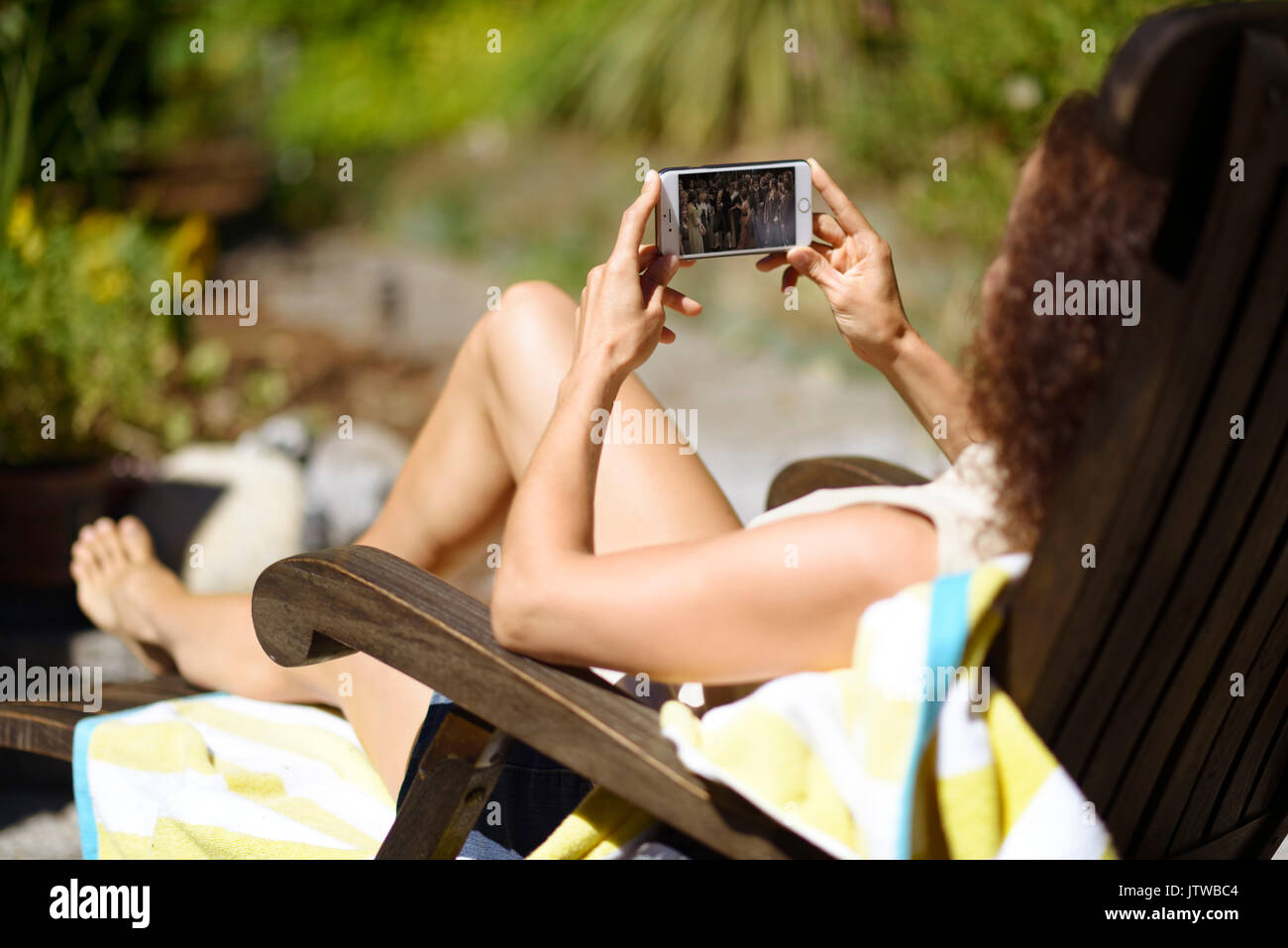 Giovane donna giacente in una sedia a sdraio al sole, a prendere il sole con un iPhone nelle sue mani, la visione di un filmato sullo schermo del telefono Foto Stock