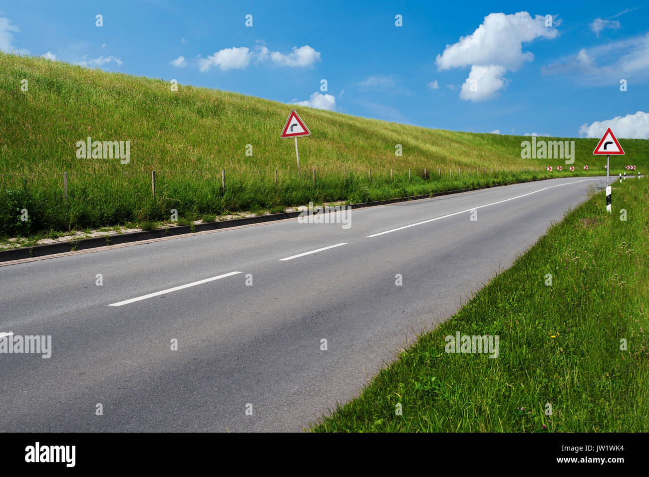 Segnale di avvertimento per indicare sharp curva a destra su entrambi i lati della strada vuota dietro una diga coperte di lussureggiante verde erba sotto il cielo di estate blu Foto Stock