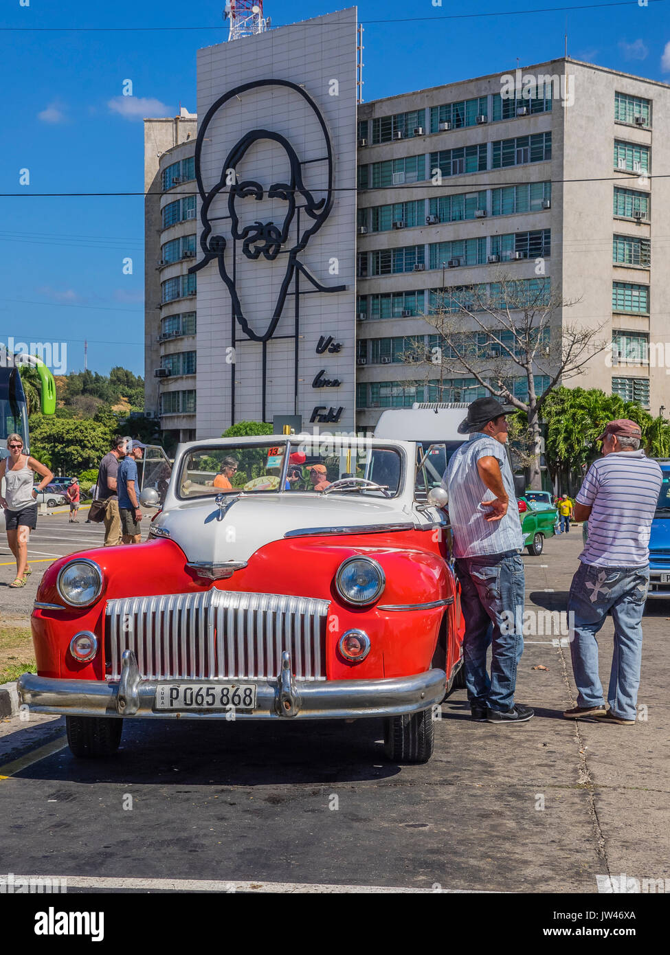 Un cubano taxi driver sorge accanto alla sua antica veicolo in Piazza della Rivoluzione in attesa per la tariffa con i turisti a piedi attorno a. Foto Stock