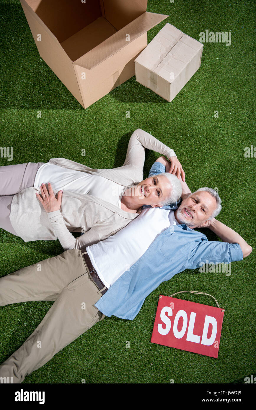 Vista superiore della felice coppia senior giacente su erba verde tra le scatole di cartone e vendute segno Foto Stock