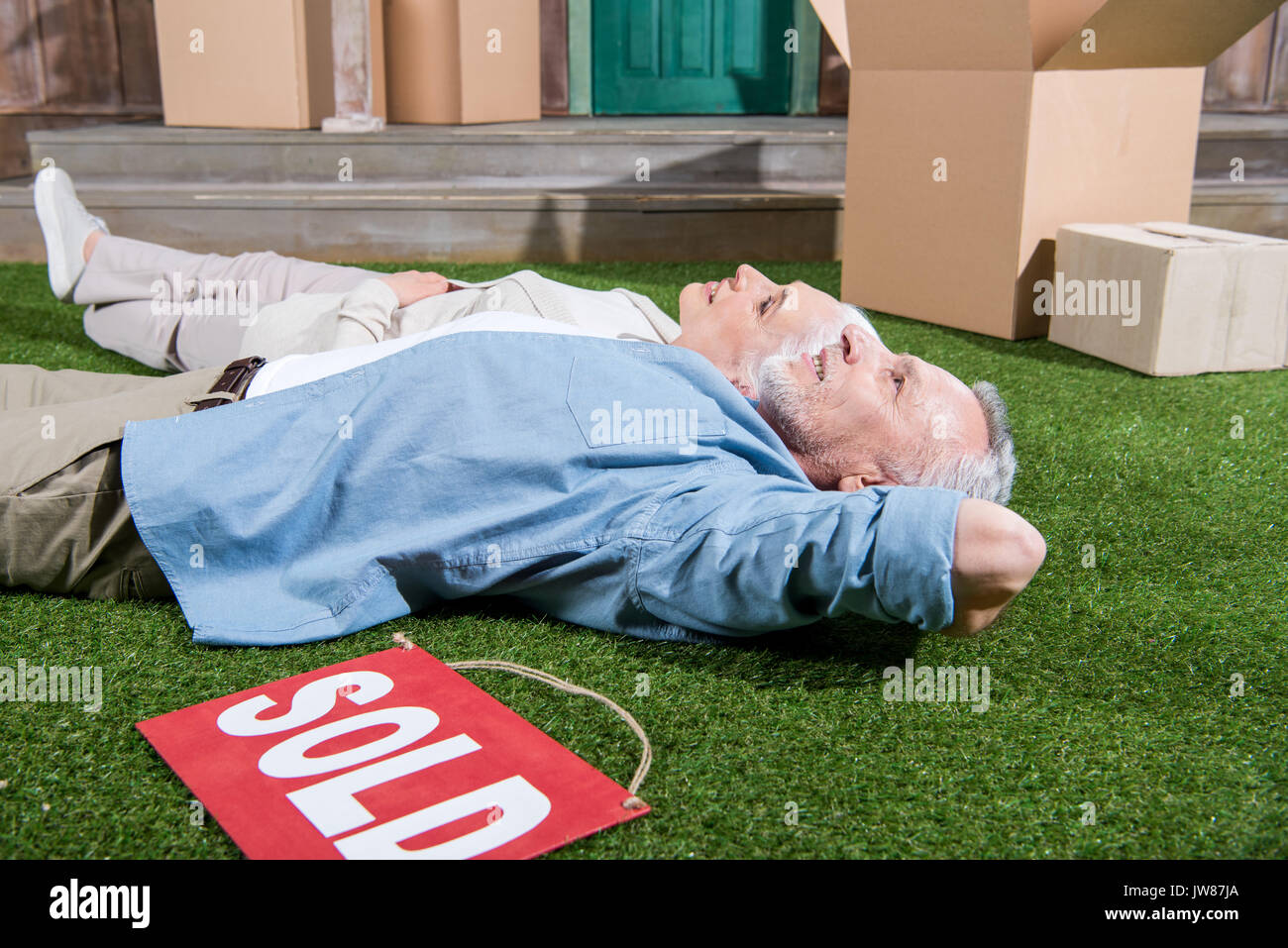 Felice coppia senior giacente su erba verde tra le scatole di cartone e vendute segno Foto Stock