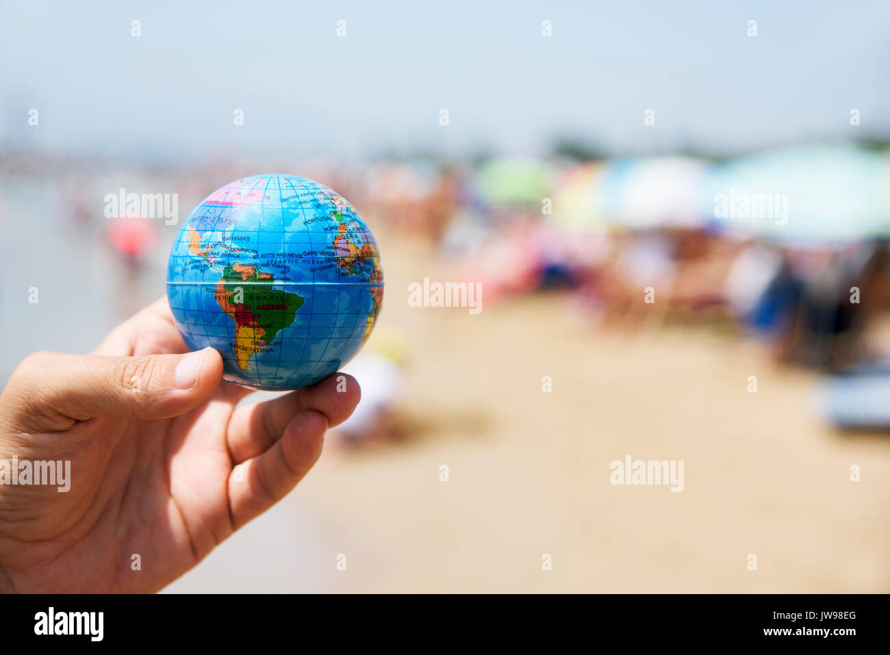 Primo piano di un giovane uomo caucasico con un globo mondo nella sua mano su una affollata spiaggia, accanto alla riva del mare Foto Stock