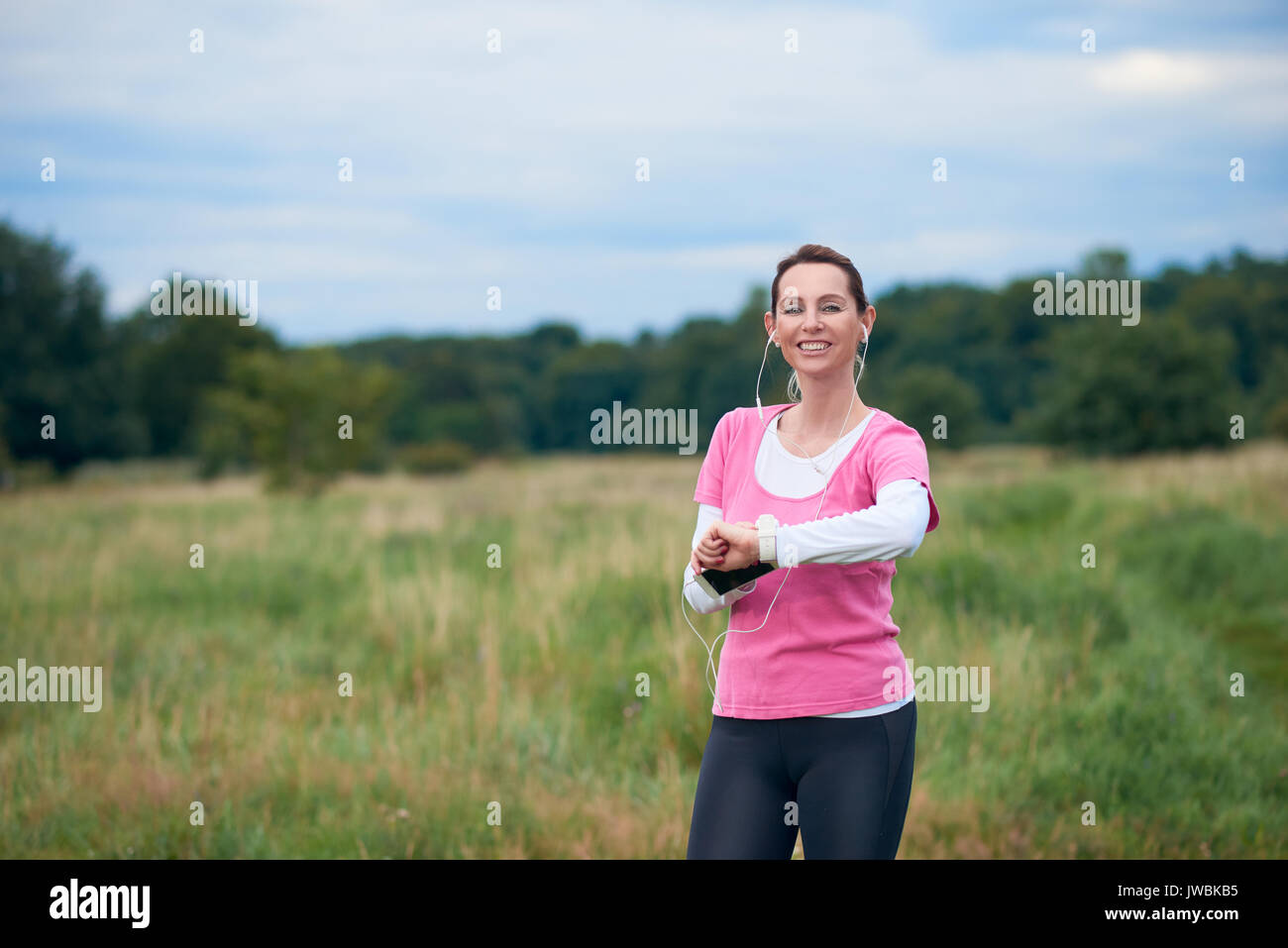 Un sano, donna sorridente nel montare l'usura con smart phone, guardare e auricolari, esercitare all'aperto in natura. Foto Stock