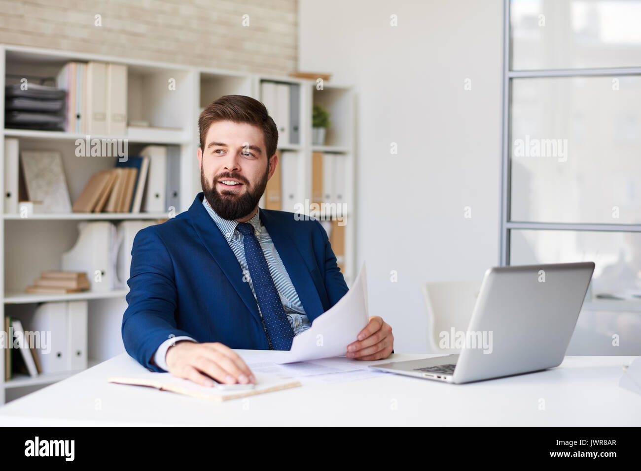 Ritratto di successo imprenditore barbuto sorridente felicemente alla ricerca di distanza mentre si lavora alla scrivania in ufficio moderno Foto Stock
