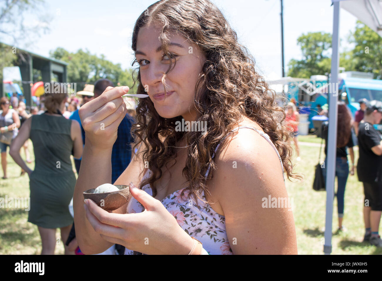 Bella giovane donna con ricci capelli castani a mangiare il gelato a Austin Gelato Festival colorati sfondo all'aperto con la folla e ombrelloni. Foto Stock