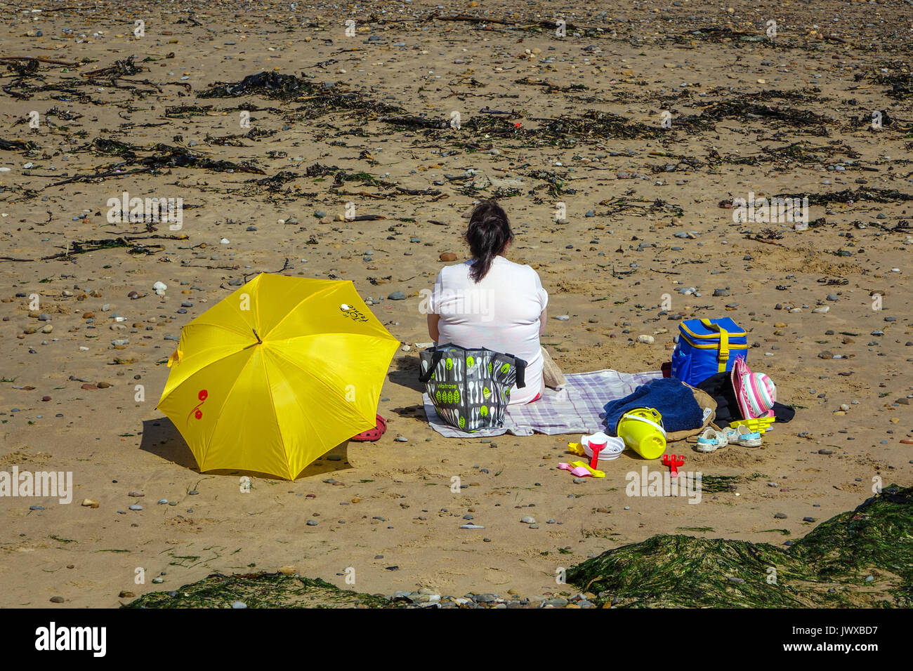 Donna sovrappeso seduti sulla spiaggia con ombrellone giallo e giocattoli di plastica, Saltburn dal mare, North Yorkshire, Inghilterra, Regno Unito Foto Stock