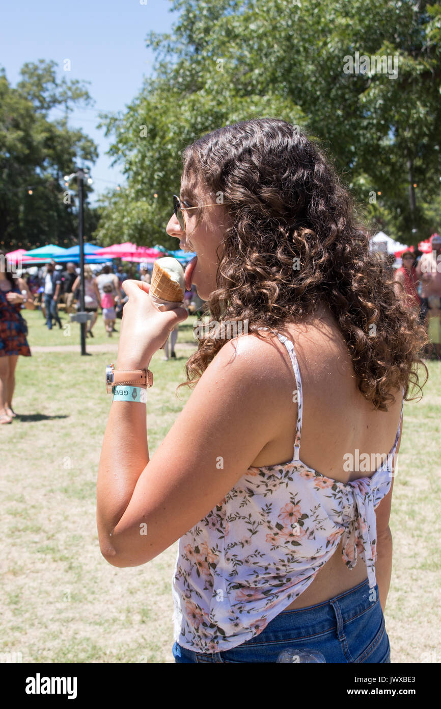 Bella giovane donna con ricci capelli castani a mangiare il gelato a Austin Gelato Festival colorati sfondo all'aperto con la folla e ombrelloni. Foto Stock