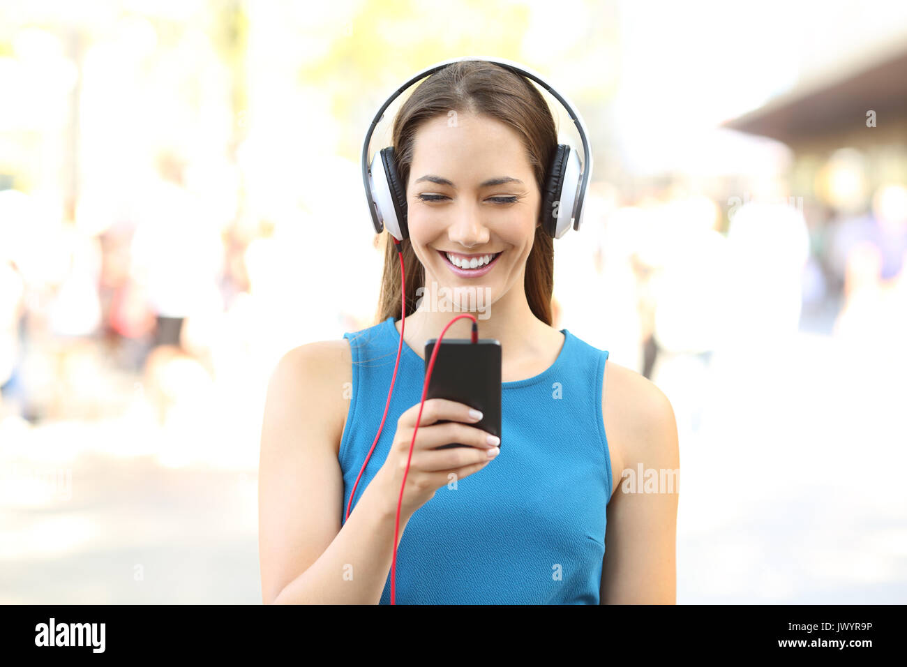 Vista frontale ritratto di una ragazza per ascoltare musica su un telefono che indossano le cuffie sulla strada Foto Stock