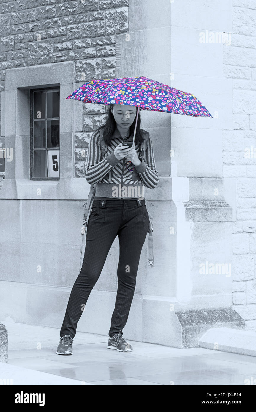 Donna che usa il telefono cellulare al riparo sotto un ombrello in una giornata piovosa bagnata a Oxford, Oxfordshire UK nel mese di agosto - monocromatico con ombrello colore schioccato Foto Stock