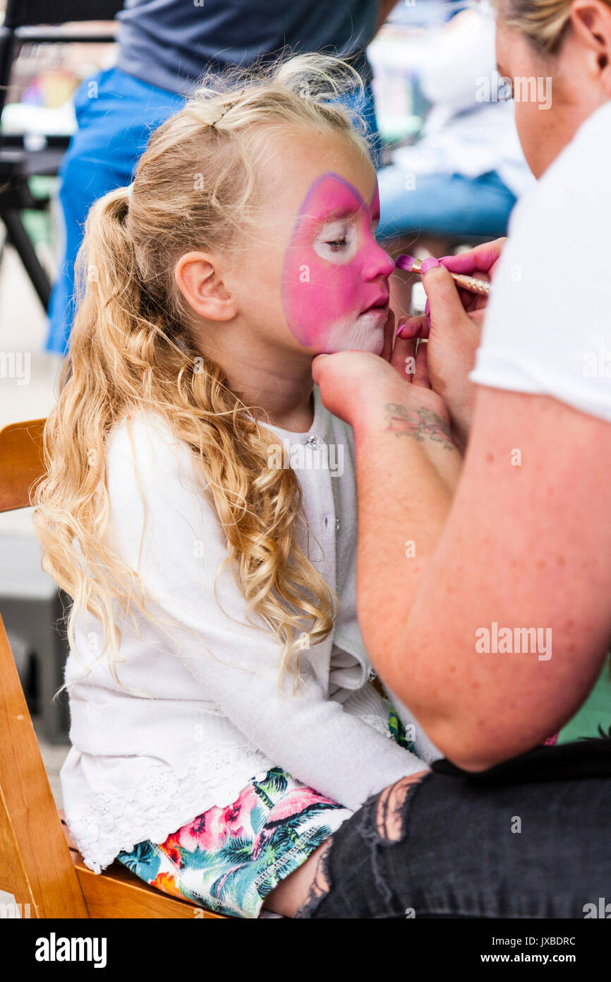 Caucasica bambino biondo, ragazza, 7-8 anni, vista laterale, avente la faccia dipinta in rosa con faccia cat. Mano che tiene il suo mento, un'altra mano azienda pennello. Foto Stock