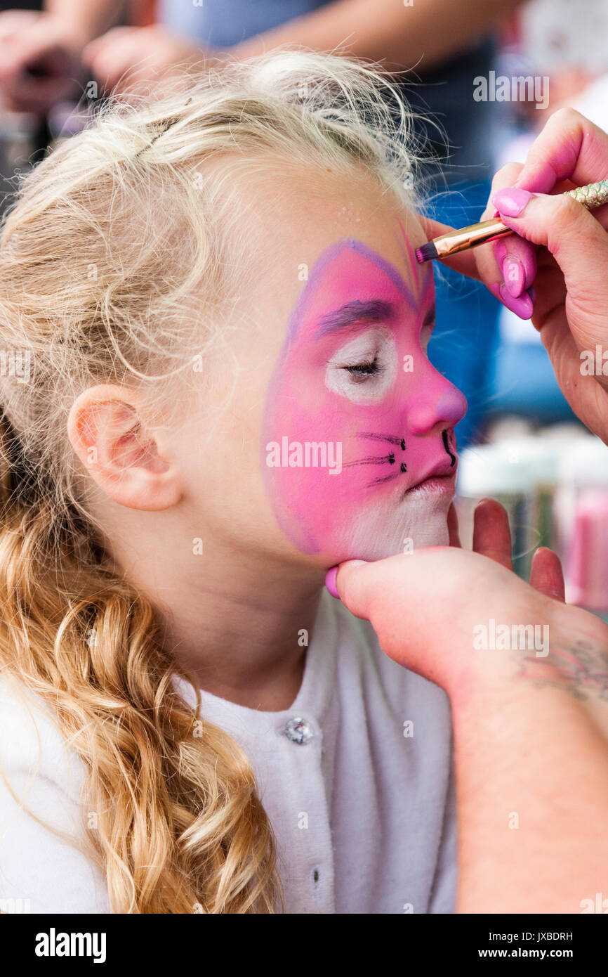 Caucasica bambino biondo, ragazza, 7-8 anni, vista laterale, avente la faccia dipinta in rosa con faccia cat. Mano che tiene il suo mento, un'altra mano azienda pennello. Foto Stock