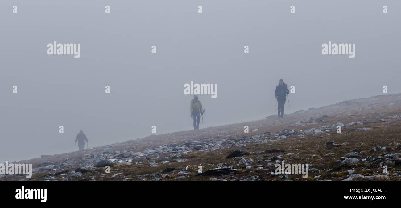 Tre i camminatori con zaini indossando indumenti di shell e walking che emergono da nebbia e pioggia mentre attraversano una montagna rocciosa ridge in th Foto Stock