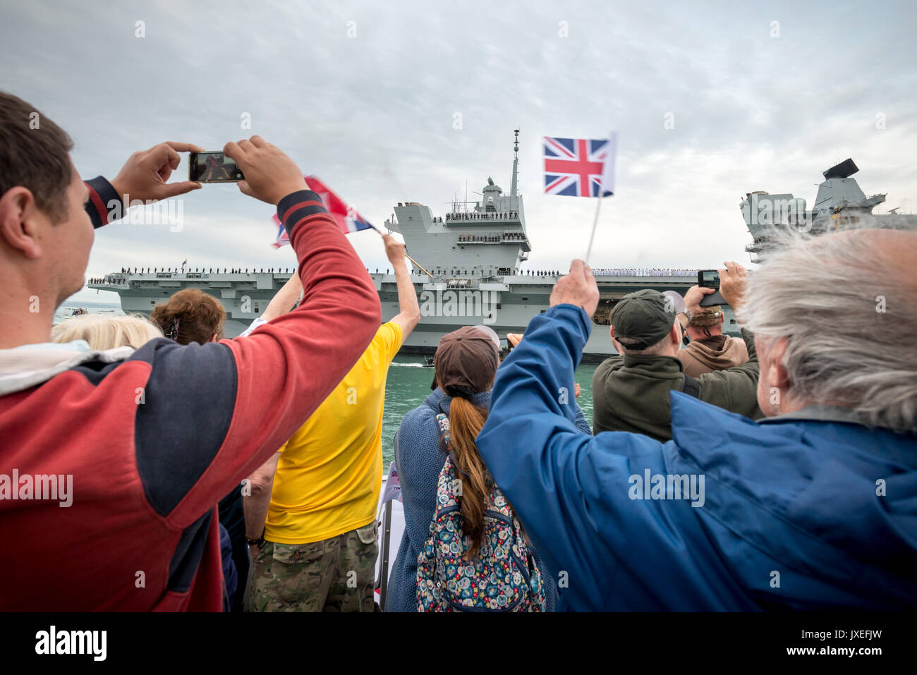 Persone ondata di bandiere e di scattare foto come si guarda l'arrivo della HMS Queen Elizabeth in Portsmouth Porto, Hampshire in Inghilterra. Foto Stock