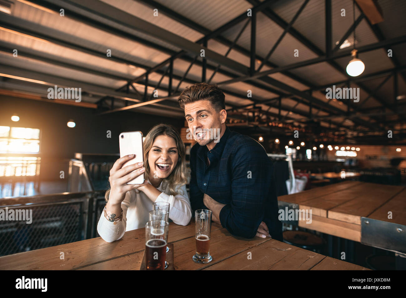 Coppia felice prendendo un selfie con uno smart phone in un bar. Uomo sorridente e la donna seduta al bar birreria con birre sul tavolo tenendo autoscatto utilizzando Foto Stock
