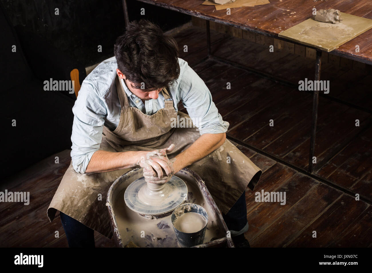 Ceramiche, workshop, arte ceramica concetto - master costituisce l'argilla sul tornio del vasaio con le mani, uomo ripiegato sul lavoro, un maschio scolpisce un arnese vicino al tavolo di legno, l'uomo in grembiule e maglietta, vista dall'alto. Foto Stock