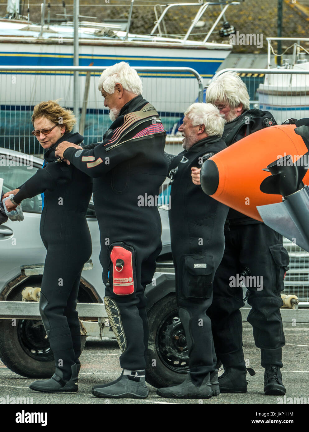 Divertente scena di quattro amici anziani si aiuta a vicenda con il montaggio del loro tute di immersione, in Penzance Harbour, Cornwall, Inghilterra, Regno Unito. Foto Stock