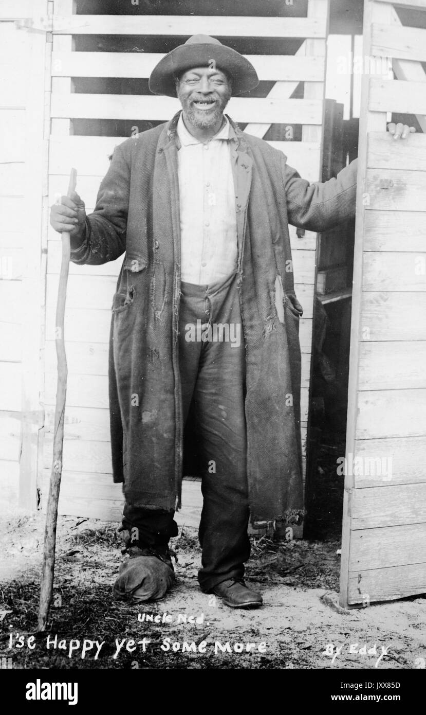 Ritratto a lunghezza intera di un uomo afroamericano maturo sorridente in piedi davanti a una serie di porte, che porta un bastone da passeggio, che indossa un cappello e un cappotto strappato, con la didascalia razzalmente pregiudiziata 'i'se Happy Yet Some More', 1915. Foto Stock