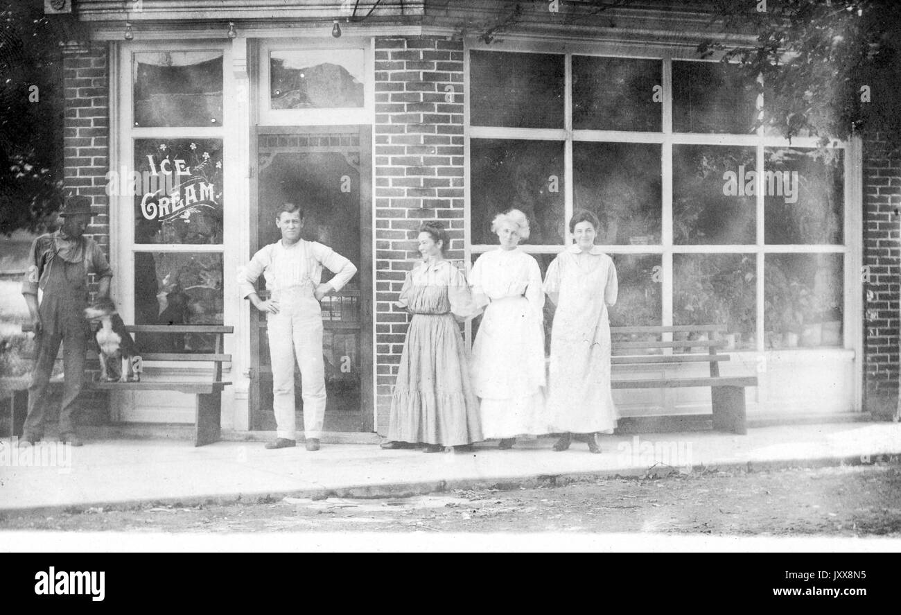 Tre donne caucasiche e un uomo caucasico maturo con espressioni felici neutrali si trovano di fronte a un fronte di magazzino che recita 'gelato', una delle donne (più a sinistra) che guarda un giovane afroamericano che sta accarezzando un cane su una panchina alla loro destra, 1915. Foto Stock