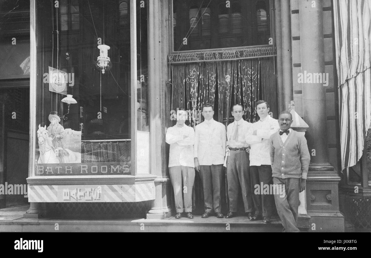 Quattro uomini caucasici maturi e un uomo afroamericano maturo, eventualmente vestito da cameriere, si trovano di fronte a un ingresso in perline a un fronte di negozi etichettato 'bagni' e 'bagni', 1915. Foto Stock