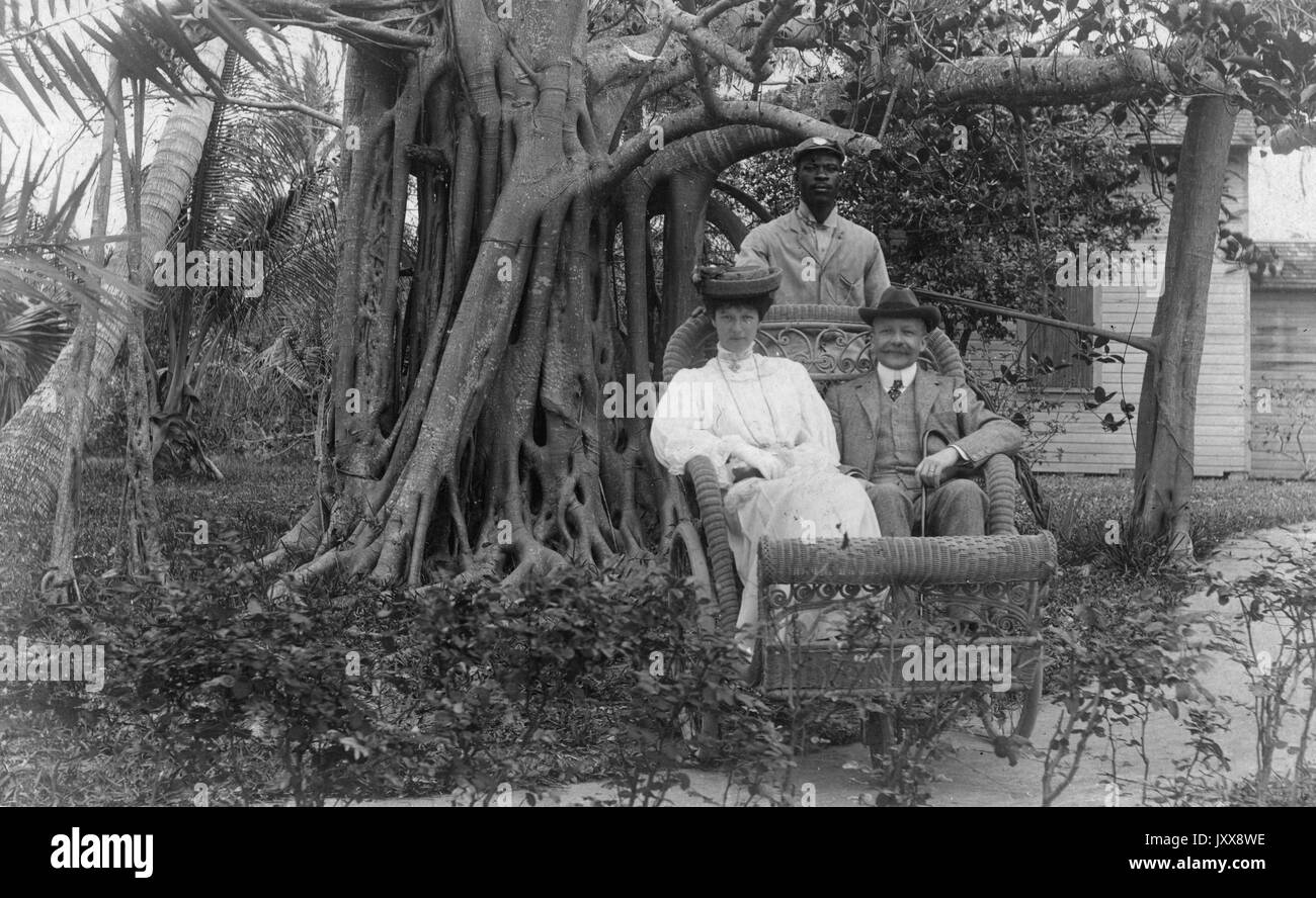 Una donna caucasica matura con un'espressione neutra e un uomo caucasico maturo sorridente siedono davanti ad alberi e piante in un carro di vimini guidato da un giovane afroamericano con un'espressione neutra, 1915. Foto Stock