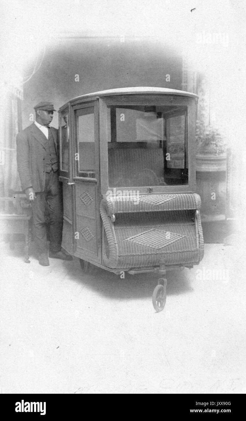 Ritratto di un uomo afroamericano maturo, un pilota di carrozza, in piedi accanto ad un carro di vimini di fronte ad uno sfondo dipinto, 1915. Foto Stock