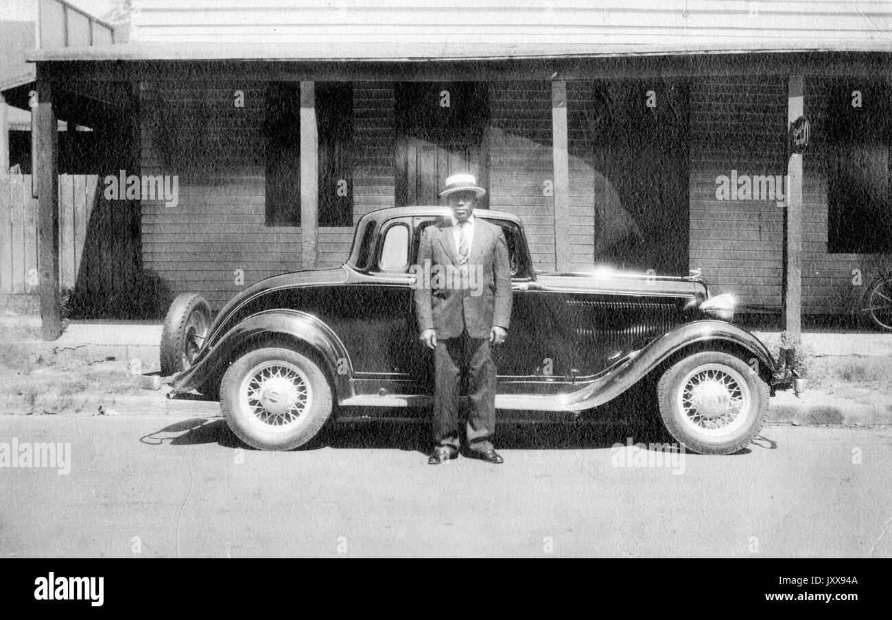 Ritratto a lunghezza intera di uomo afro-americano in piedi davanti ad un'automobile all'aperto, indossando un vestito e un cappello, espressione facciale neutra, 1920. Foto Stock