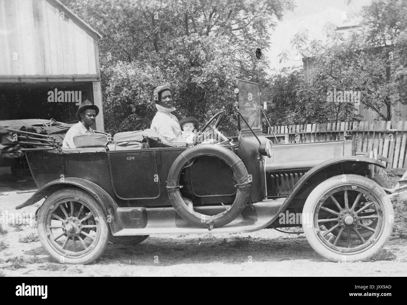Foto di paesaggio a lunghezza intera di due uomini afroamericani maturi in un'automobile, un uomo nella parte posteriore, un uomo al volante, un bambino caucasico nella parte anteriore, all'aperto, espressioni facciali neutre, 1920. Foto Stock