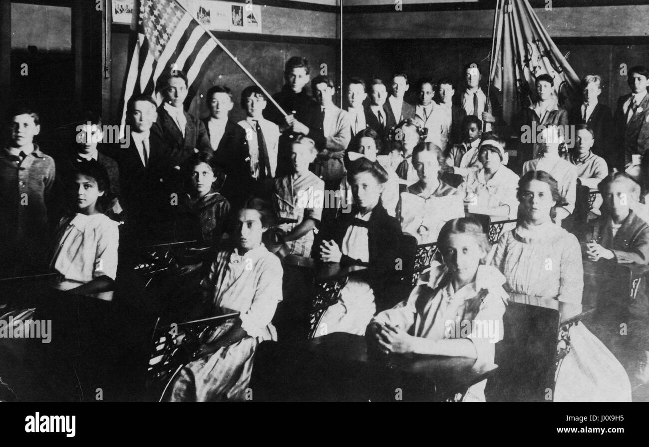 Paesaggio full length shot di studenti caucasici al chiuso seduti a scrivanie, espressioni facciali neutrali, uno studente che tiene una bandiera americana, uno studente afroamericano seduto nella schiena, 1920. Foto Stock