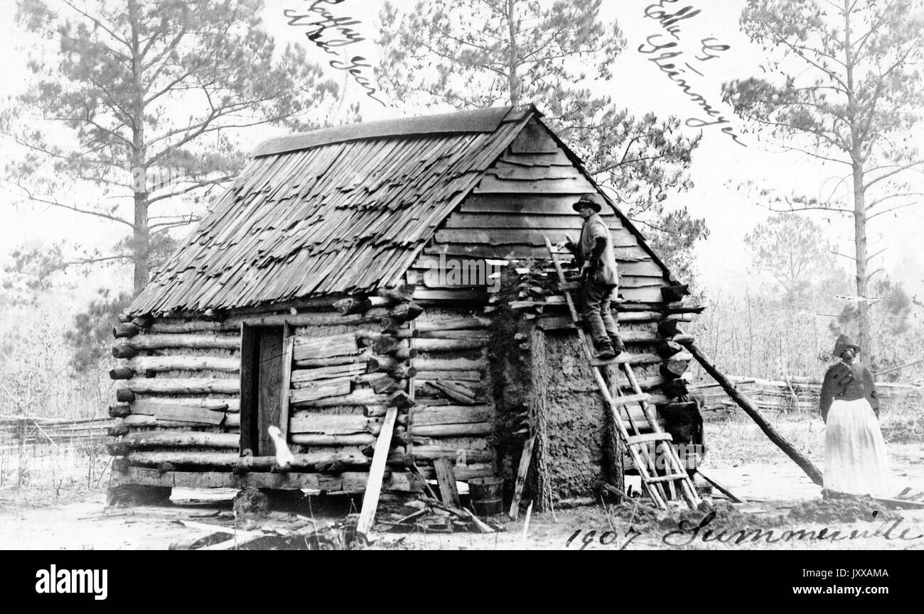 Ritratto a lunghezza intera di uomo e donna afro-americani davanti alla cabina di legno, giacca leggera da uomo, pantaloni e cappello scuri, donna che indossa una blusa scura, gonna e cappello chiari, maschio sulla scala in piedi accanto alla cabina, alberi sullo sfondo, espressioni neutre, 1915. Foto Stock