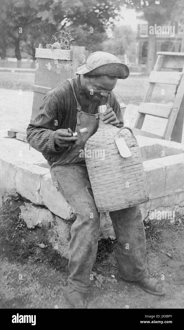 Ritratto seduta di un maschio afroamericano maturo che indossa una camicia scura, tute e cappellino, tenendo e guardando una caraffa tessuta, seduto su una struttura in cemento davanti a casa e alberi, espressione sorridente, 1915. Foto Stock