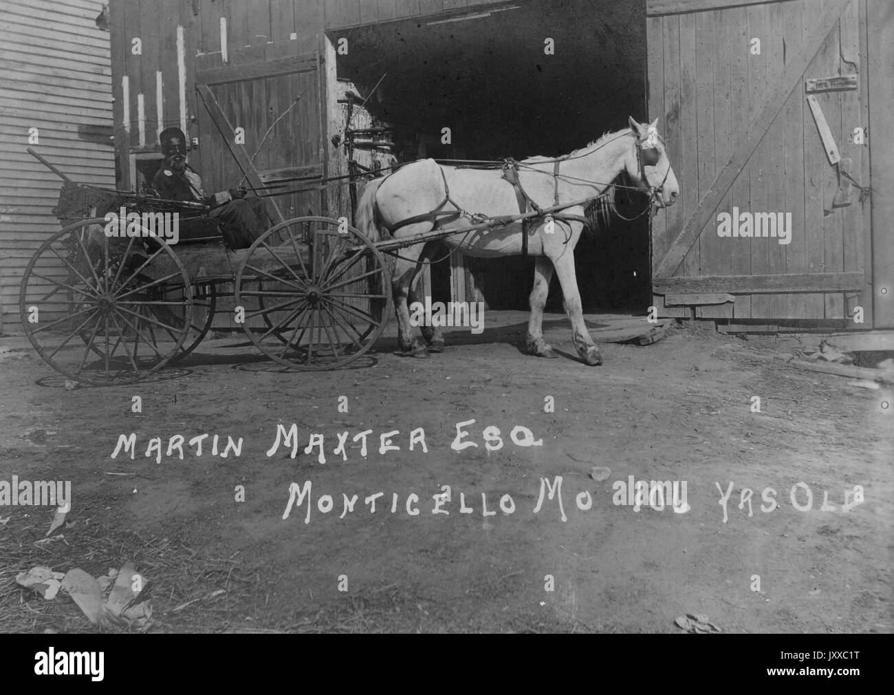 Ritratto a lunghezza intera di un uomo afro-americano in buggy guidato da cavallo, con abiti scuri, seduto accanto alla porta aperta del fienile, espressione neutra, 1915. Foto Stock