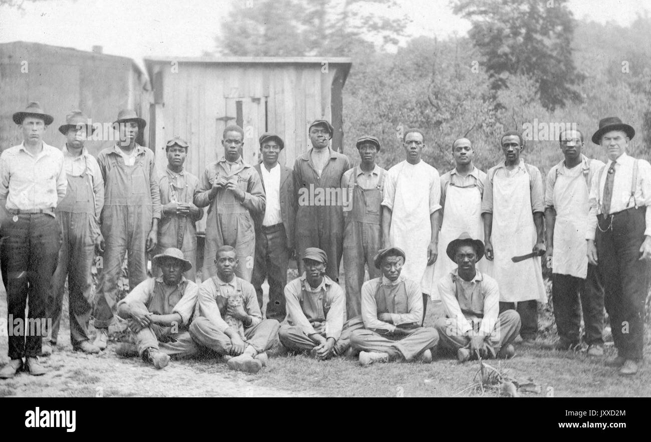 Un gruppo di giovani e maturi lavoratori afroamericani con espressioni neutre si trovano fuori da edifici di baraccopoli, con due uomini caucasici (all'estrema sinistra e all'estrema destra) vestiti con abiti più raffinati, che sembrano essere i loro superiori, 1915. Foto Stock