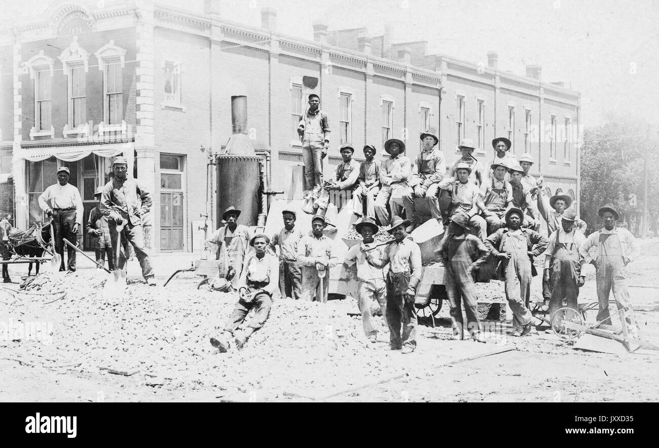 Foto di paesaggio a lunghezza intera di un gruppo di lavoratori afro-americani che indossano cappelli e uniformi, la maggior parte sono seduti su alcuni macchinari, pochi in piedi, 1915. Foto Stock