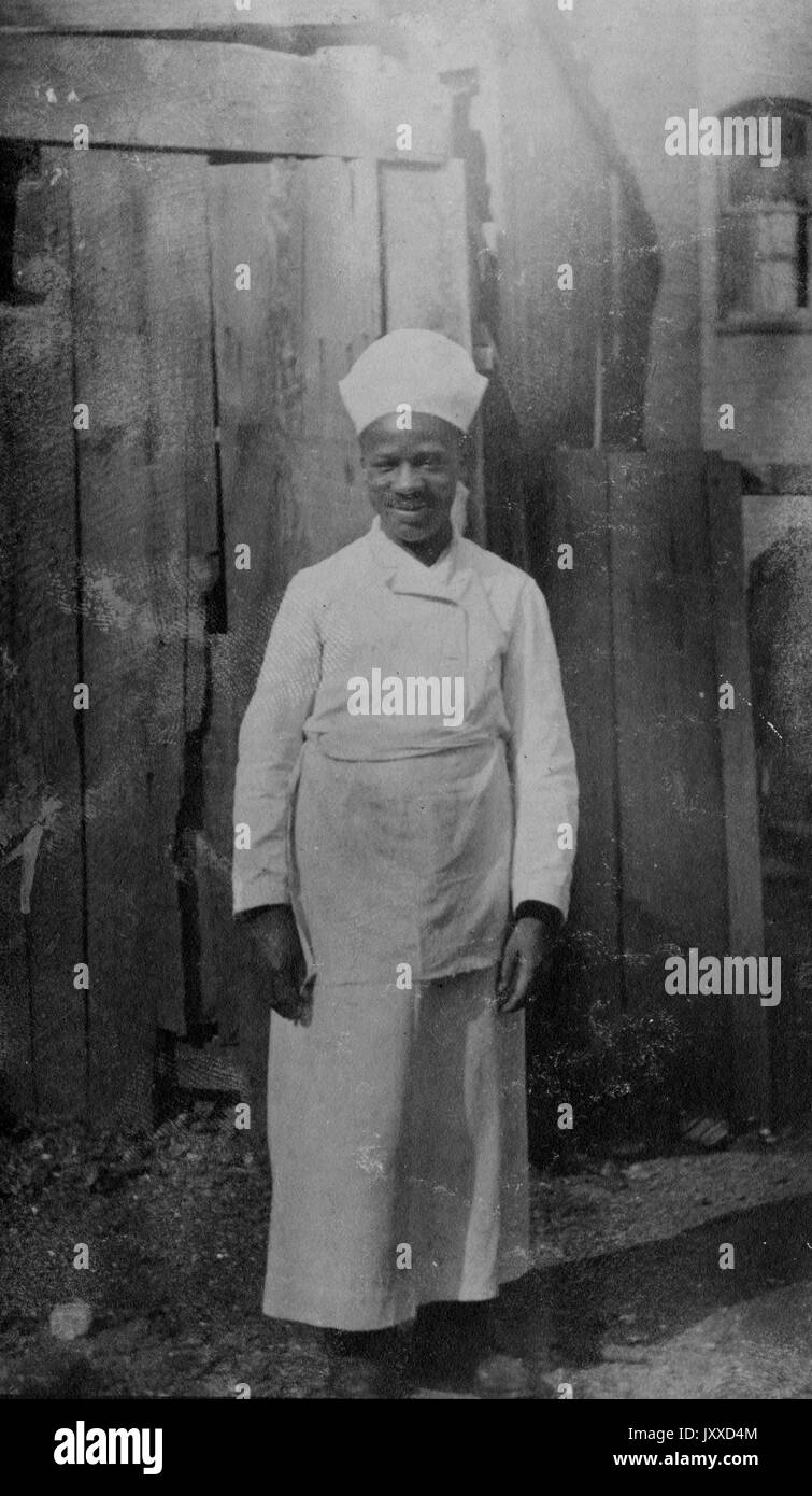 Ritratto a lunghezza intera di un uomo afro-americano, vestito in grembiule e cappellino di uno chef o di un marinaio, in cucina, sorridente espressione facciale, 1920. Foto Stock