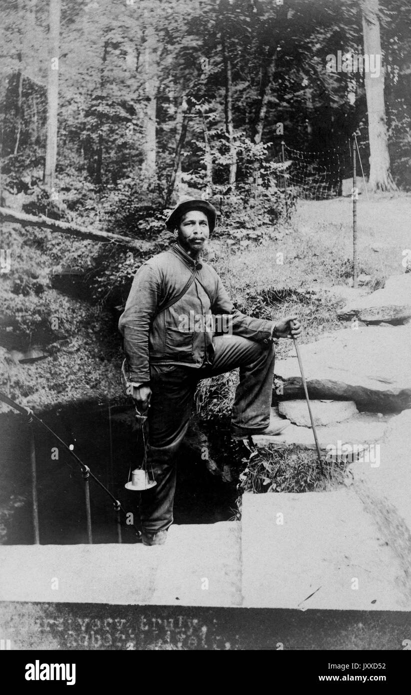 A piena lunghezza Ritratto di afro-americano all'aperto tenendo una lanterna e un bastone da passeggio, vestito in abiti campestre, folle espressione facciale, 1920. Foto Stock